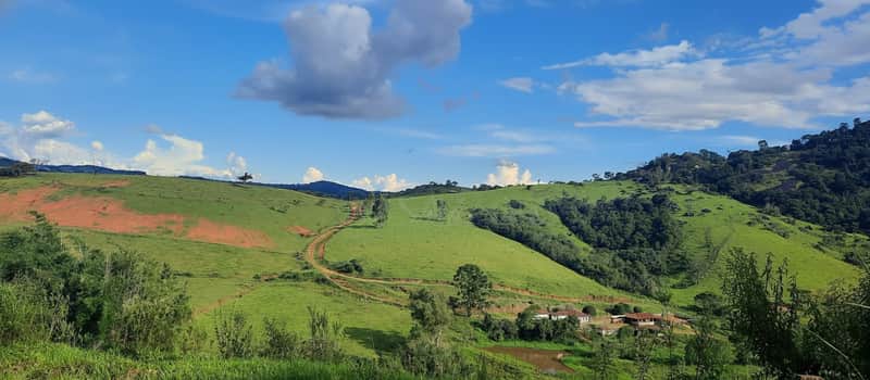Paisagem rural ensolarada com morros verdes, estrada de terra e casas ao fundo. Céu azul com nuvens.