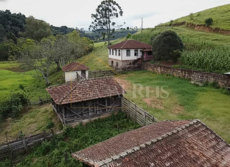 Área externa de fazenda com casa antiga e construções rústicas. Telhados de cerâmica e pasto verde.