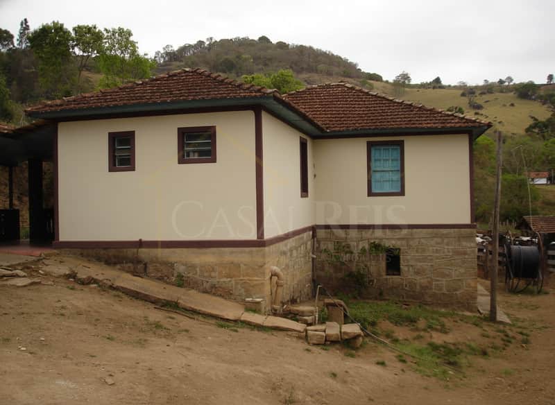 Fachada de casa rústica com paredes claras, base em pedra e janelas coloridas em área externa campestre.