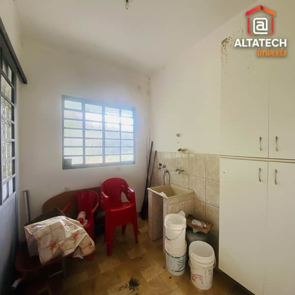 Área de serviço com pia, armário branco, janela gradeada e cadeiras de plástico vermelhas.