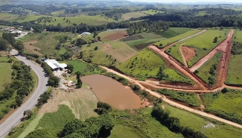 Vista aérea de área rural com lago, estrada e construções. Paisagem verde com plantações e morros ao fundo.