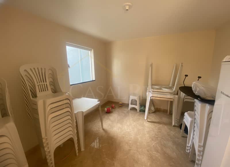 Sala clara com piso brilhante, mesa e cadeiras de plástico brancas empilhadas. Janela com luz natural.