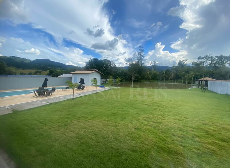 Área externa com piscina, gramado, lago e montanhas ao fundo em dia ensolarado com nuvens.