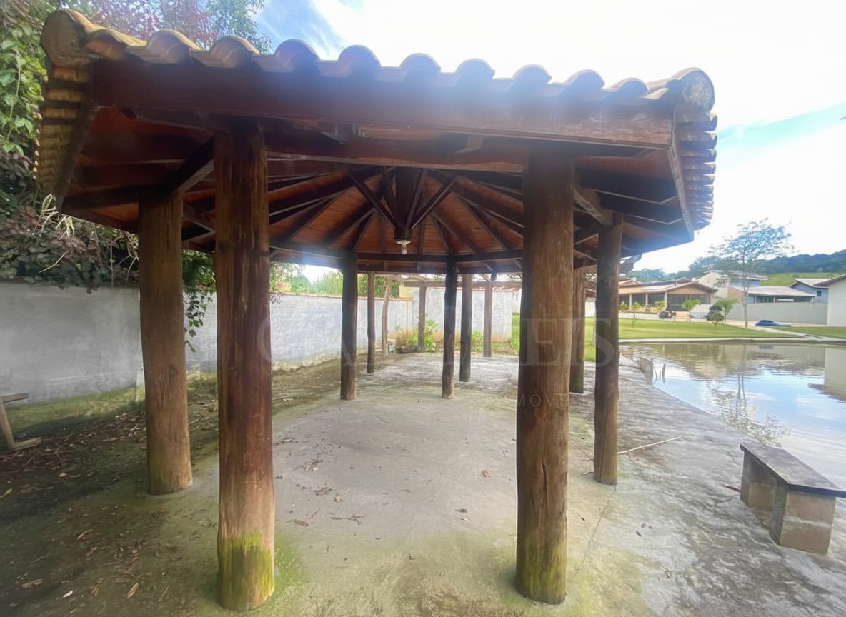 Área externa com quiosque rústico de madeira, piso de concreto e lago com banco.