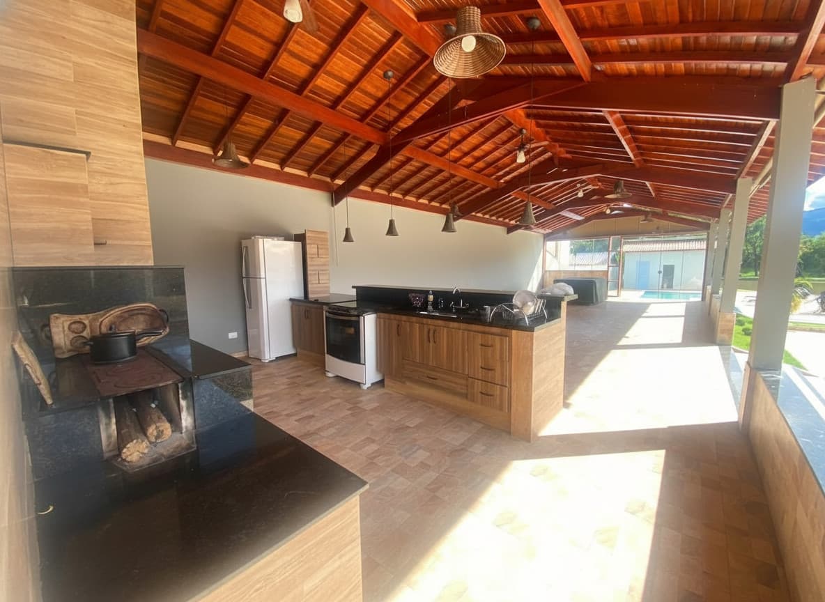 Área externa gourmet com churrasqueira, fogão a lenha, armários de madeira e piscina ao fundo.