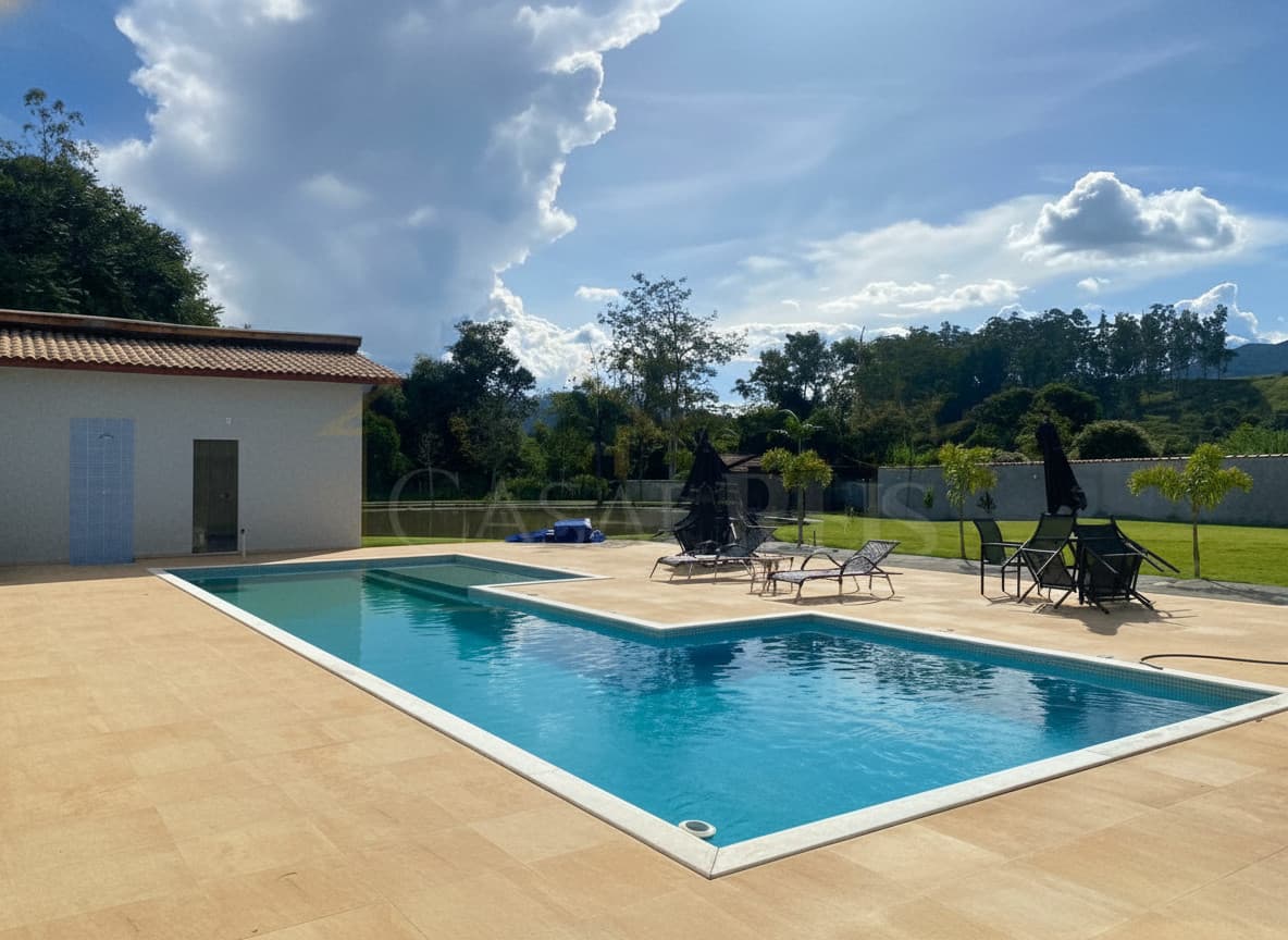 Área externa com piscina azul e espreguiçadeiras em deck bege, sob céu azul com nuvens.