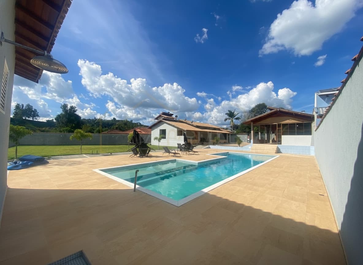 Área externa com piscina azul, espreguiçadeiras e gramado verde sob céu azul com nuvens.