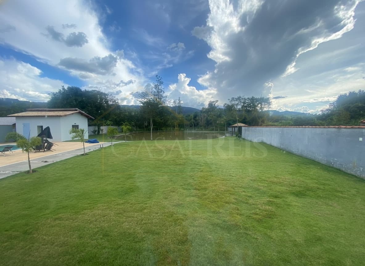 Ampla área externa com gramado, lago e construção. Céu azul com nuvens.